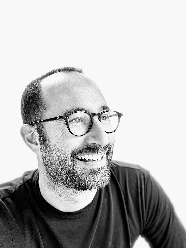 Black and white portrait of Ross Barna in a dark T-shirt and round glasses, laughing and looking off to the side.